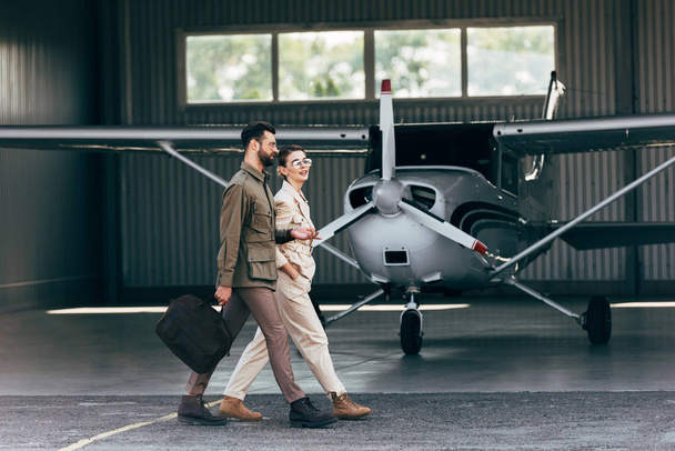 vista lateral do homem levando saco e andando com namorada elegante perto do hangar com avião
  - Foto, Imagem