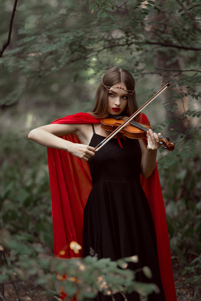 schöne mystische Mädchen in rotem Mantel spielt auf Geige in dunklen Wäldern - Foto, Bild