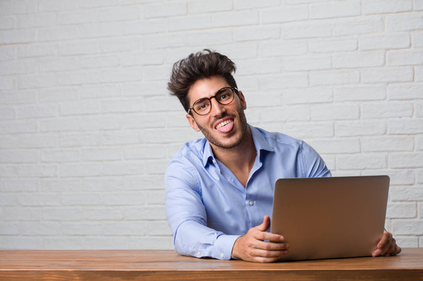Молодий бізнесмен сидить і працює на ноутбуці вираження впевненості і емоцій, весело і дружньо, показуючи язик як знак гри або веселощів
 - Фото, зображення