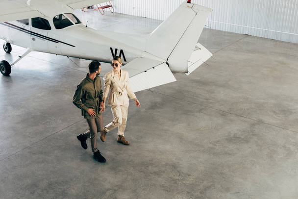 high angle view of young couple in stylish jackets walking in hangar with airplane  - Foto, immagini