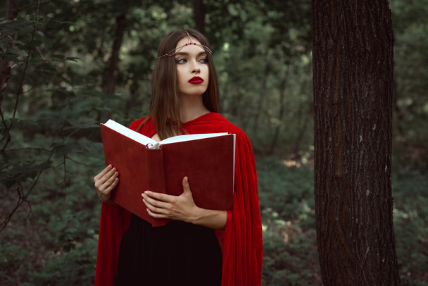 elegantes mystisches Mädchen in rotem Mantel mit magischem Buch im Wald - Foto, Bild