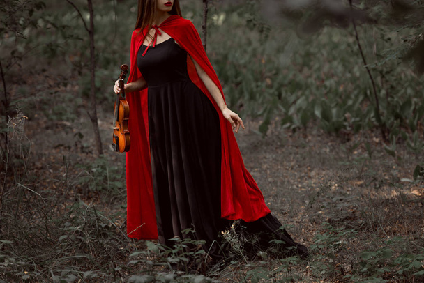 περικοπεί άποψη του κομψή γυναίκα μυστηριακό μαύρο φόρεμα και κόκκινο μανδύα, κρατώντας βιολί στο δάσος - Φωτογραφία, εικόνα
