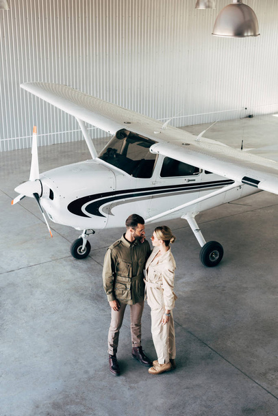 visão de alto ângulo de casal em jaquetas elegantes olhando um para o outro no hangar com avião
  - Foto, Imagem