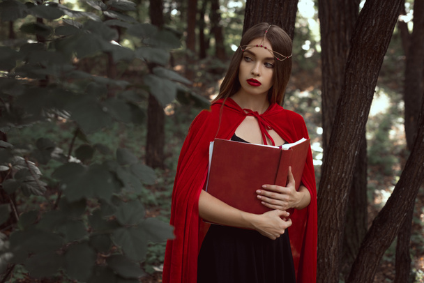 attractive girl in red cloak with magic book in dark woods - Foto, imagen