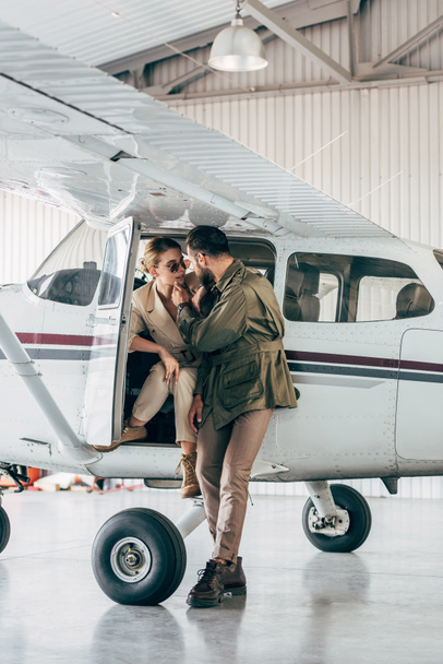 young fashionable couple in jackets looking at each other near airplane  - Фото, изображение