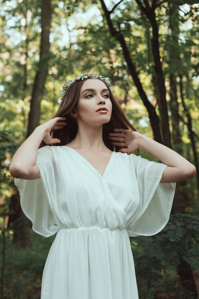 young tender woman posing in white dress and floral wreath in forest - Foto, Imagem