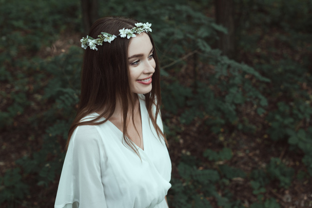 stylish smiling girl posing in white dress and floral wreath in woods - Photo, image