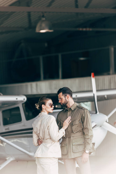  fashionable young couple in stylish jackets standing in hangar with airplane  - Foto, Imagem