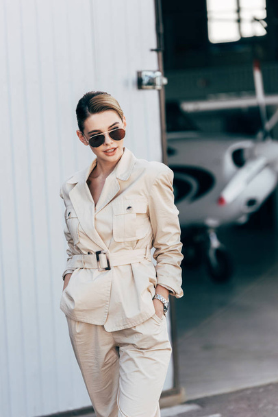 mooie jongedame in zonnebrillen en jas poseren met handen in de zakken in de buurt van vliegtuig  - Foto, afbeelding