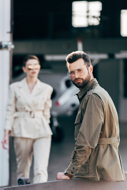 selective focus of man in eyeglasses and jacket looking at camera while his girlfriend walking near hangar with plane - Φωτογραφία, εικόνα