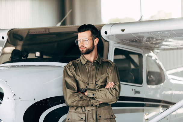 young man in green jacket and eyeglasses posing with crossed arms near airplane  - Photo, Image