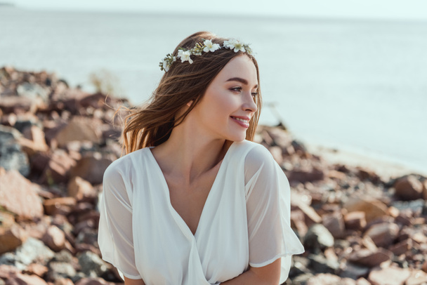 attractive smiling girl posing in floral wreath on rocky beach near sea - Photo, image