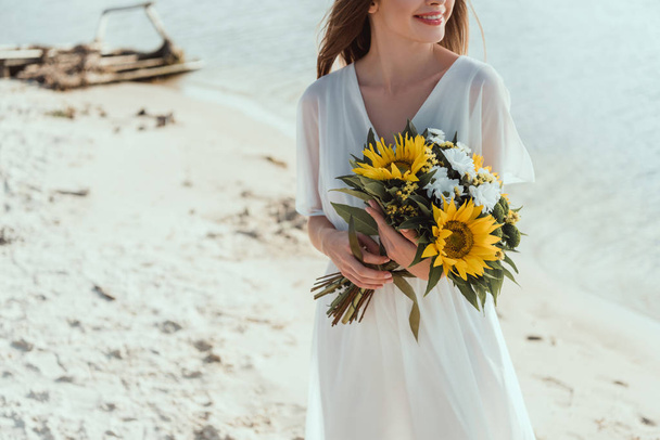 cropped view of smiling girl holding bouquet with sunflowers on beach - Фото, изображение