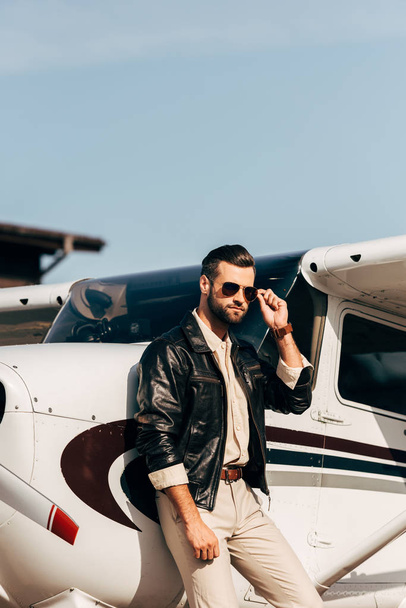 уверенный в себе пилот в кожаной куртке и солнцезащитных очках позирует возле самолета
 - Фото, изображение