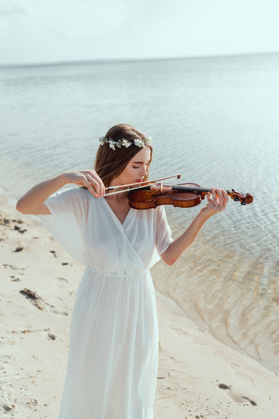 beautiful woman in elegant white dress and floral wreath playing violin on seashore - Фото, зображення