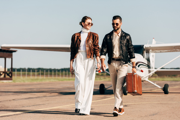 feliz jovem casal em jaquetas de couro e óculos de sol andando com mala retro perto do avião
  - Foto, Imagem
