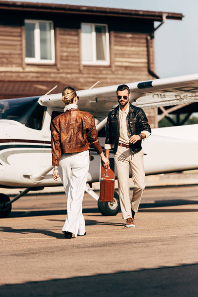 stylish woman in leather jacket giving vintage suitcase to boyfriend near airplane  - Zdjęcie, obraz