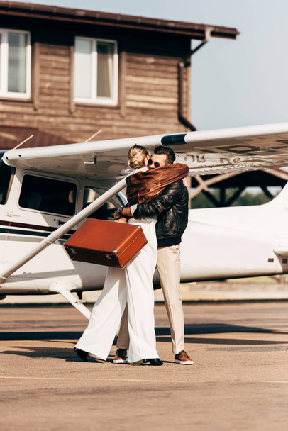 young couple in leather jackets with vintage suitcase hugging each other near airplane  - Photo, Image