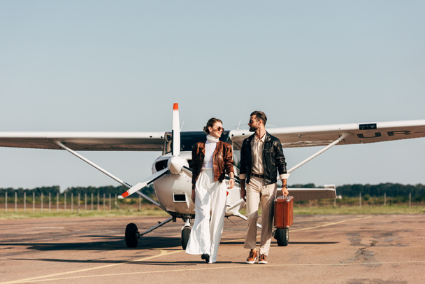 κομψό νεαρό ζευγάρι στο δερμάτινο μπουφάν και γυαλιά ηλίου, περπάτημα με ρετρό βαλίτσα κοντά σε αεροπλάνο  - Φωτογραφία, εικόνα