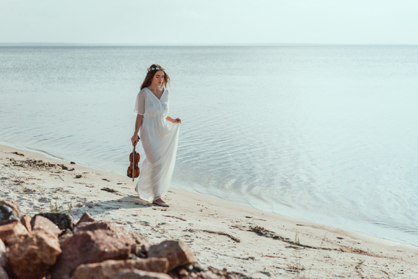 attractive young woman in elegant dress with violin walking on beach near sea - 写真・画像