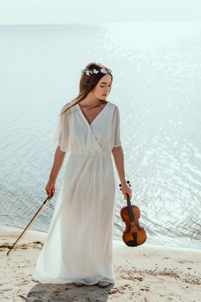 beautiful elegant woman in dress and floral wreath holding violin on beach near sea - Фото, изображение