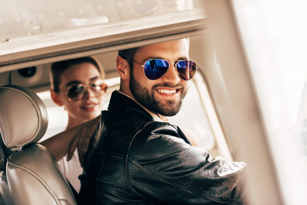 piloto varón feliz en gafas de sol y chaqueta de cuero mirando a la cámara mientras su novia sentada cerca en cabina de avión
  - Foto, imagen