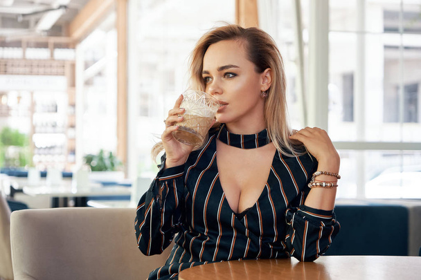 Uma mulher jovem e bem sucedida tomando uma bebida sentada em uma mesa em um café moderno enquanto espera seu parceiro. Blusa sensual com decolagem, rosto bonito e pele. Mãos segurando um belo copo
. - Foto, Imagem
