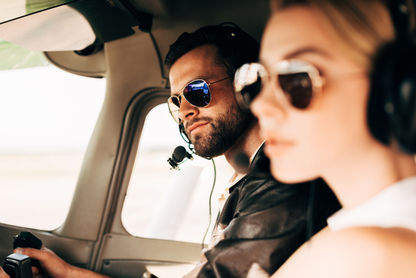 enfoque selectivo de piloto masculino guapo en auriculares y gafas de sol sentado con novia en cabina de avión
 - Foto, Imagen