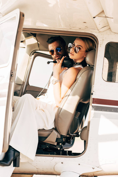 atractiva mujer joven en gafas de sol y auriculares sobre el cuello sentado cerca de novio en cabina de avión
  - Foto, imagen