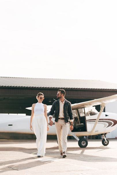 joven pareja de moda en gafas de sol caminando cerca de avión
  - Foto, imagen