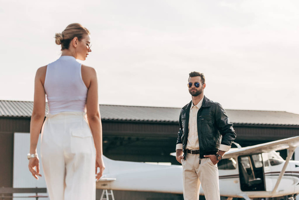 luottavainen nuori mies nahkatakki ja aurinkolasit katselee tyttöystävä lähellä lentokonetta
  - Valokuva, kuva
