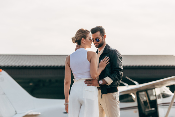 hombre con estilo en chaqueta de cuero y gafas de sol abrazando novia atractiva cerca de avión
 - Foto, imagen