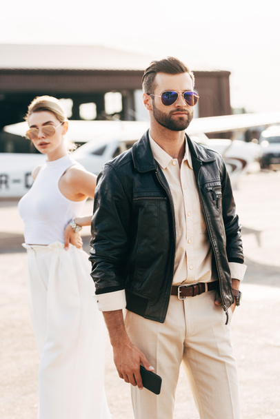красивый стильный мужчина в солнцезащитных очках и кожаной куртке, держащий смартфон, в то время как его девушка стоит рядом с самолетом
  - Фото, изображение