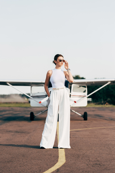 atractiva mujer joven con estilo en gafas de sol mirando hacia otro lado y posando cerca de avión
 - Foto, Imagen