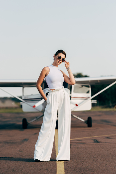 κομψό νεαρή γυναίκα σε γυαλιά ηλίου θέτοντας κοντά σε αεροπλάνο - Φωτογραφία, εικόνα