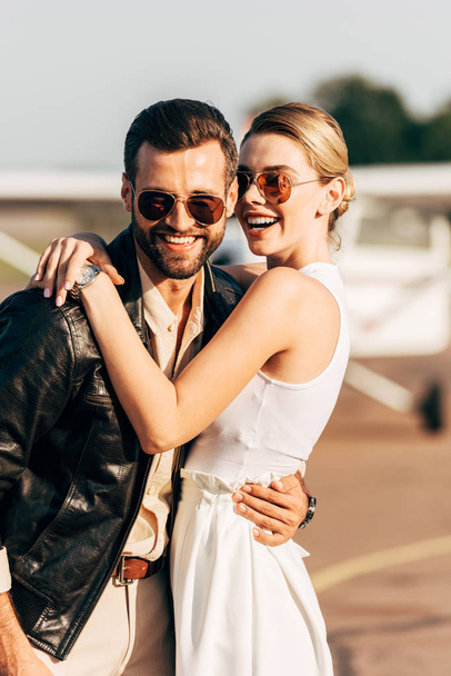 lachende attraktive Frau umarmt stilvollen Freund in Lederjacke und Sonnenbrille in der Nähe von Flugzeug  - Foto, Bild