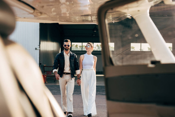 enfoque selectivo de hombre con estilo en chaqueta de cuero y gafas de sol caminando con novia cerca de avión
 - Foto, imagen