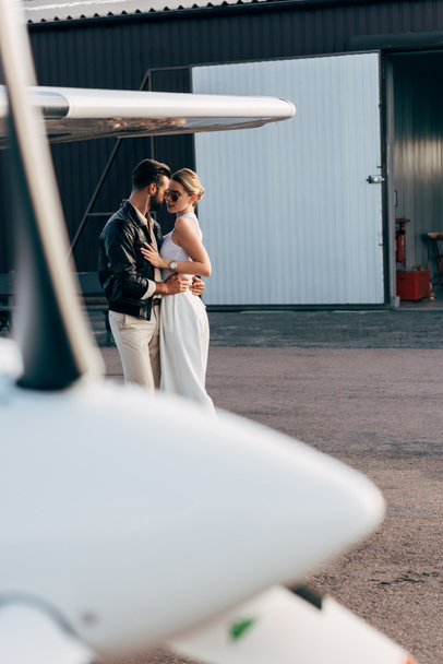 man in leather jacket and sunglasses embracing attractive girlfriend near plane - Fotó, kép