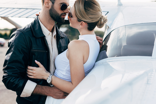 schöner junger Mann in Lederjacke und Sonnenbrille umarmt Freundin in Flugzeugnähe - Foto, Bild