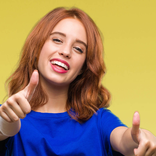 Young beautiful woman over isolated background approving doing positive gesture with hand, thumbs up smiling and happy for success. Looking at the camera, winner gesture. - Photo, Image