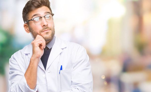 Young handsome man wearing doctor, scientis coat over isolated background with hand on chin thinking about question, pensive expression. Smiling with thoughtful face. Doubt concept. - Foto, immagini