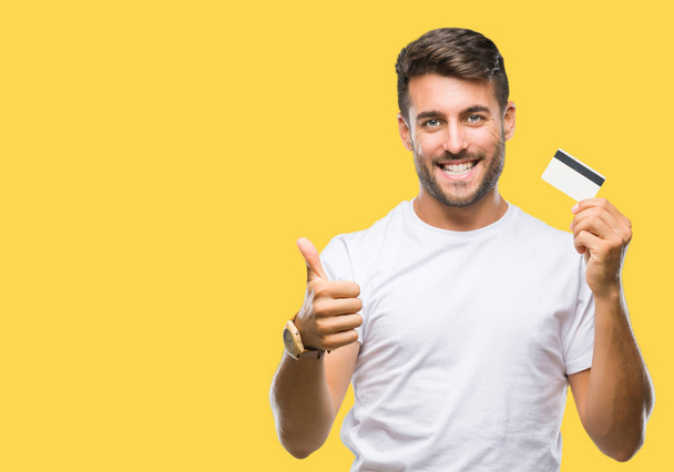 Young handsome man holding credit card over isolated background happy with big smile doing ok sign, thumb up with fingers, excellent sign - Photo, Image