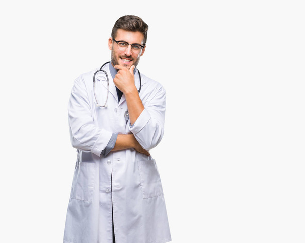 Młody przystojny lekarz mężczyzna na białym tle patrząc pewny siebie na aparat z uśmiechem z skrzyżowanymi rękami i ręką uniesioną do góry na podbródek. Myślenie pozytywne. - Zdjęcie, obraz
