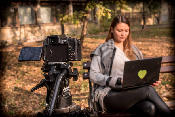 Videokamera kuvaa nuorta naista istumassa penkillä puistossa, jossa on sylimittari
  - Valokuva, kuva