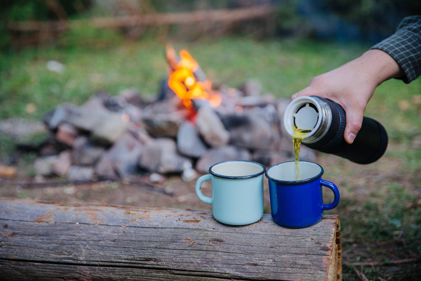 hand pour tea into metal cups. camp fire on background. hiking concept - Photo, image