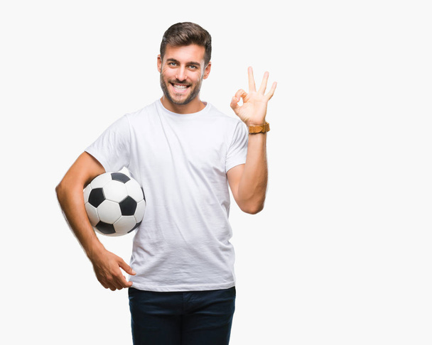 Όμορφος νεαρός, κρατώντας την μπάλα ποδοσφαίρου ποδοσφαίρου πάνω από απομονωμένες φόντο κάνει εντάξει πινακίδα με τα δάχτυλα, εξαιρετική σύμβολο - Φωτογραφία, εικόνα
