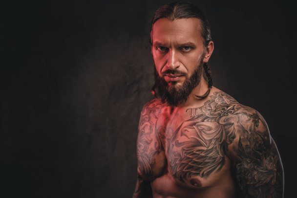 Foto de cerca de un brutal hombre tatuado sin camisa y barbudo. Aislado sobre un fondo de textura oscura
. - Foto, Imagen