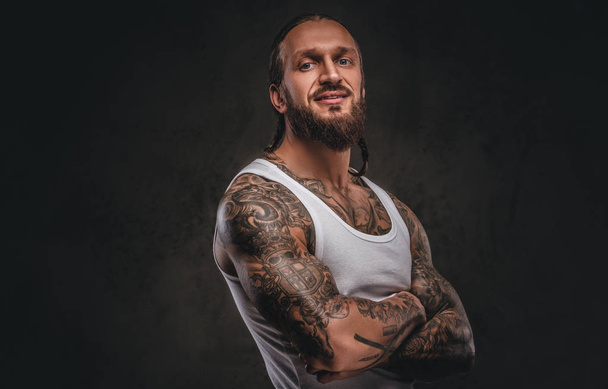 Brutal bearded tattooed male in white shirt posing with crossed arms. Isolated on a dark textured background. - Photo, Image