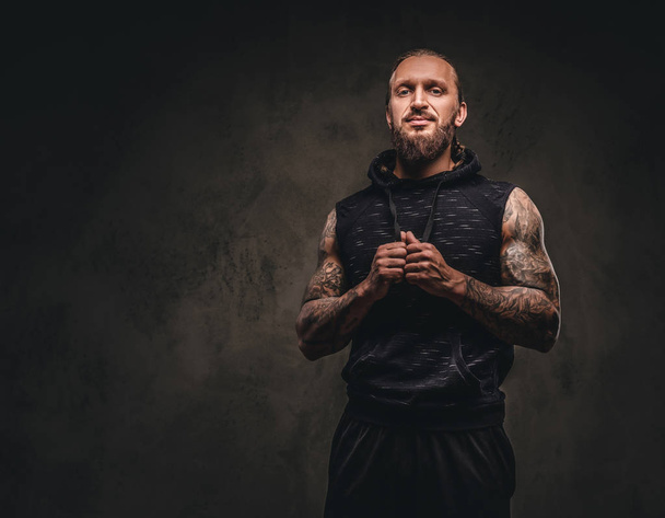 Portret van een bebaarde getatoeëerd atleet in een zwarte hoodie. Geïsoleerd op een donkere gestructureerde achtergrond. - Foto, afbeelding