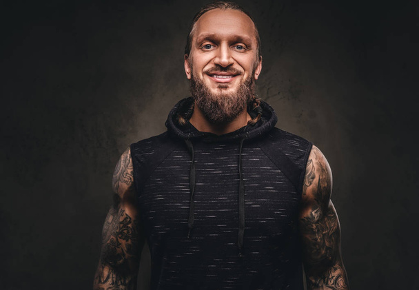 Retrato de un atleta tatuado barbudo con capucha negra. Aislado sobre un fondo de textura oscura
. - Foto, imagen
